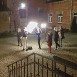 Fotografia przedstawiająca dziedziniec wydziału nocą. Widać na niej osoby studenckie przygotowujące plan oświetleniowy w plenerze.