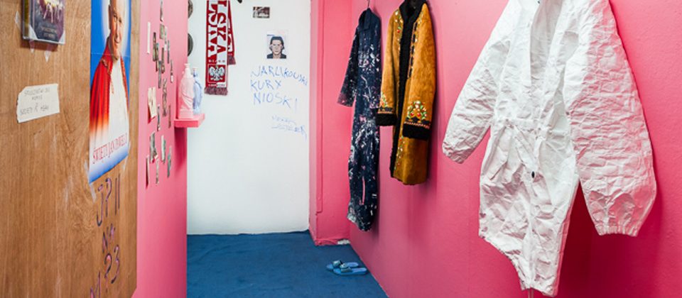 fotografia przedstawiająca korytarz z niebieska podłogą i różowymi ścianami na których na wieszakach wiszą ubrania w różnych kolorach