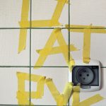 plakat przedstawiający fragment ściany z białymi płytkami ceramicznymi i kontaktem. Na płytkach żółtą papierową taśmą wyklejone imię Patryk.