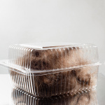 Fotografia kolorowa przedstawiająca plastikowe przezroczyste pudełko spożywcze wypełnione brązowymi ściętymi włosami. Pudełko leży na lustrzanym blacie. Tło zdjęcia jest jasnoszare.