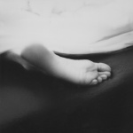 Fotografia czarno-biała przedstawiająca stopę na czarnym tle wystającą spod białej tkaniny.Palce stopy skierowane są w stronę prawego dolnego rogu zdjęcia a pięta w stronę lewego górnego rogu.