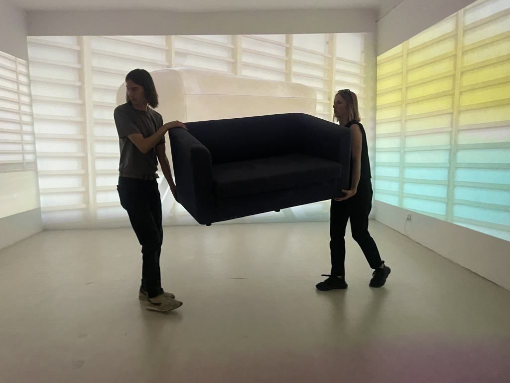 Fotografia przedstawiająca dwoje ludzi przenoszących sofę w pustym wnętrzu. Na białych ścianach widać projekcje drabinek gimnastycznych.