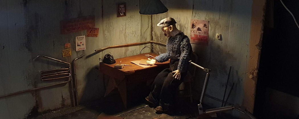 Fotografia przedstawiająca makietę do animacji poklatkowej. W makiecie widoczny jest starszy człowiek siedzący przy biurku w ciemnym pokoju rozświetlonym jedynie światłem lampy nad biurkiem.